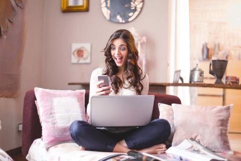 Girl sits cross-legged on coach with laptop on her legs but looking excitedly at her smartphone