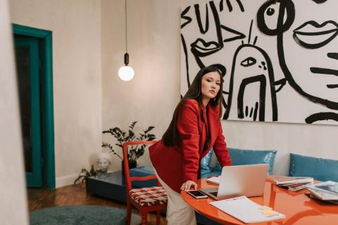 A girl in a red blazer leans at a table with a laptop and phone on it and abstract art on the wall